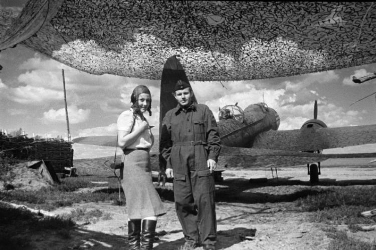 Это фото было сделано известным фотокором Совинформбюро Семеном Фридляндом на аэродроме в Липицах. На снимке: Оксана Фридлянд, супруга фотографа, и авиамеханик полка. На заднем фоне можно увидеть бомбардировщик Ил-4. 